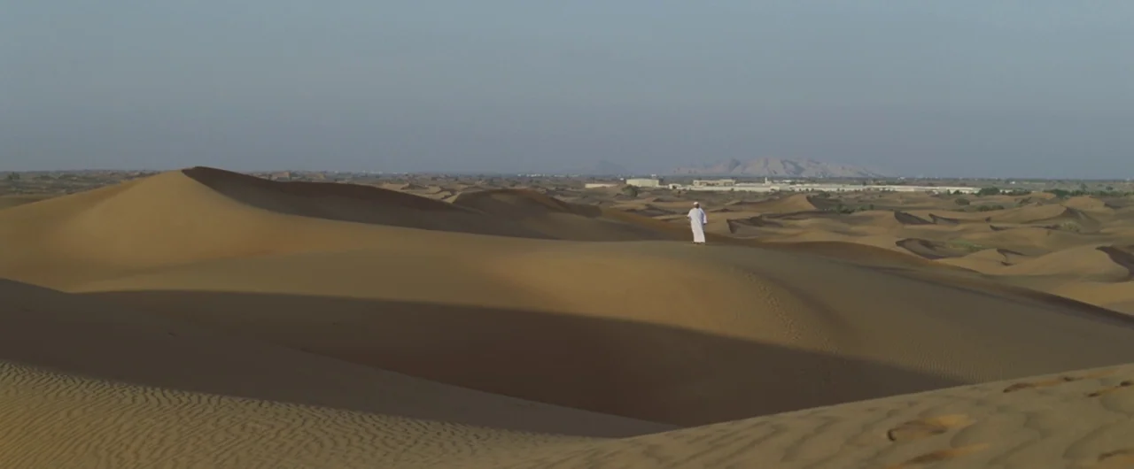 Syriana (2005) movie still: wide — A vast, rolling desert landscape features deep gold sand dunes under a hazy sky. A single man in a white thobe…