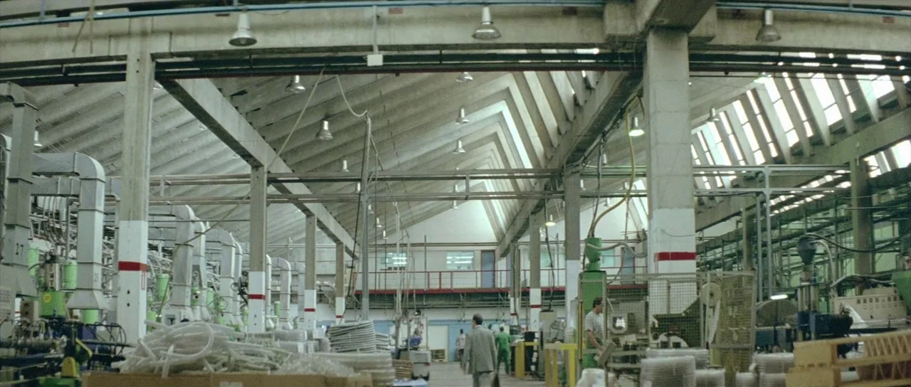 Dogtooth movie still: wide — A large industrial factory features high ceilings with exposed metal trusses and skylights. Several workers are…