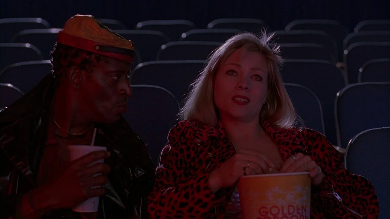 Whore (1991) movie still: medium — Two people sit in a dim movie theater with rows of empty dark seats behind. The woman wears a leopard print…