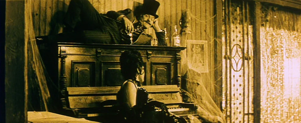 Lemonade Joe (1964) movie still: wide — An interior saloon featuring a wooden piano, cobwebs, and a beaded curtain entryway. A man reclines on top…