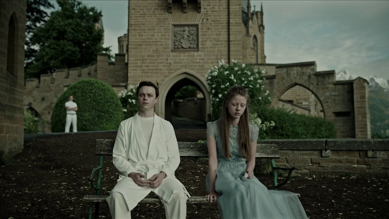 A Cure for Wellness (2017) movie still: fullbody — A stone castle facade with a large arched entryway looms behind a park bench. A young man and…