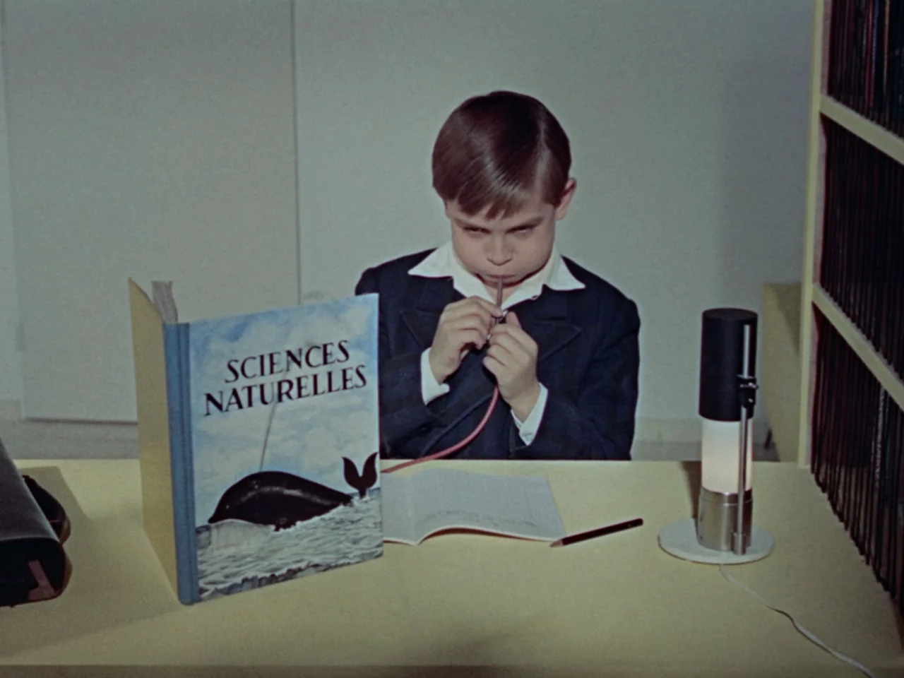 Mon Oncle (1958) movie still: medium — A young boy sits at a yellow desk in a room with white walls. He focuses intently on blowing through a thin…