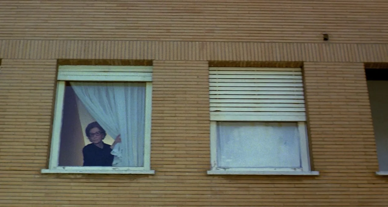 Tenebre (1982) movie still: wide — A flat brick building facade features multiple windows with varied curtain and blind states. A woman stands…