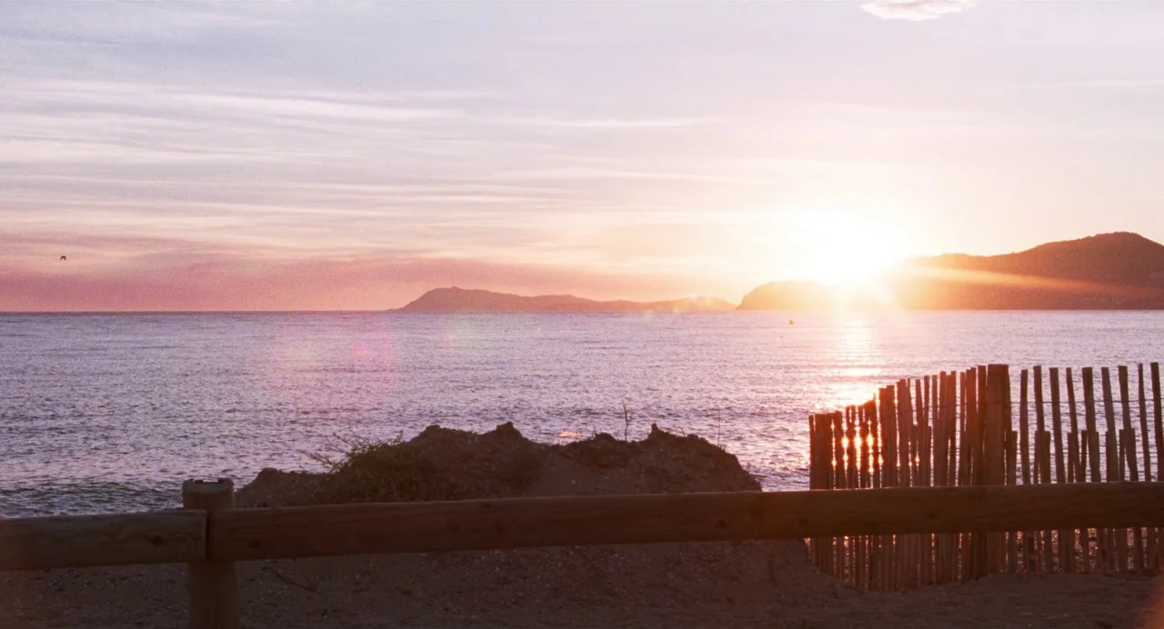 Godard Mon Amour (2015) movie still: establishing — A calm sea landscape at sunset with rolling hills in the distant background. A wooden…