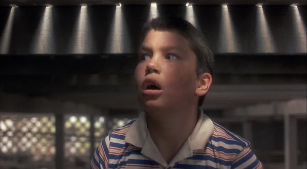 Stand By Me (1986) movie still: closeup — A dark, cramped wooden area with light filtering down through floor joists. A young boy looks toward the…