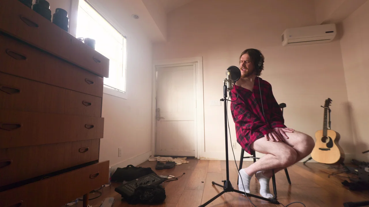 Bo Burnham: Inside (2021) movie still: medium — A spare room contains a tall chest of drawers, an acoustic guitar, and equipment. A bearded man in a…