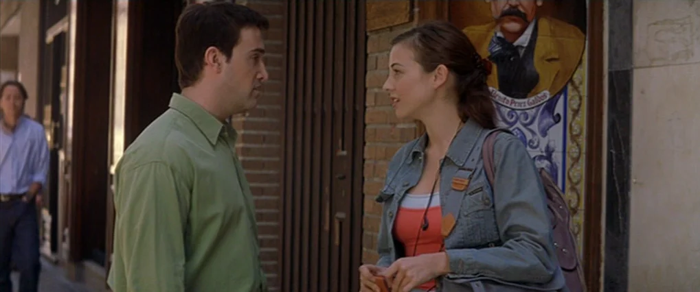 Talk To Her (2002) movie still: medium — An urban sidewalk with a brick wall and a decorative tiled sign. A man and a woman stand close to each…