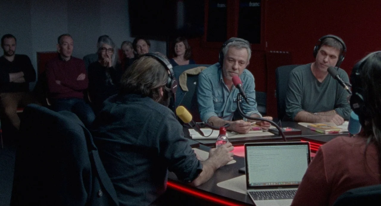 Non-Fiction (2018) movie still: medium — A dimly lit room with a long table occupied by several people in microphones. Three men participate in a…