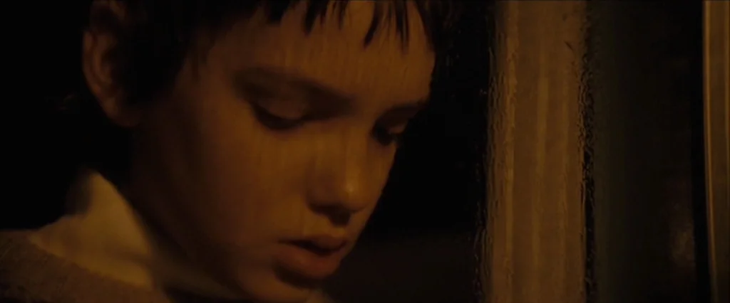 Let Me In (2010) movie still: closeup — A close view of a teenage boy pressed against a dark window at night. The dim light highlights his facial…
