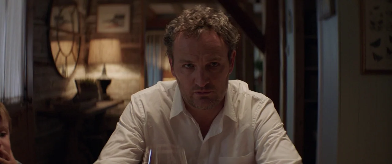 Pet Sematary (2019) movie still: medium — A dark dining room with wooden walls and a blurred table lamp background. A middle-aged man sits centered…