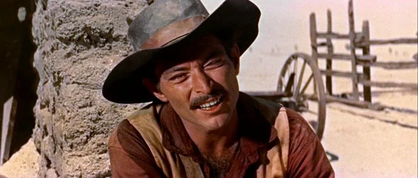 Ride Lonesome (1959) movie still: medium — A man in a worn hat stands against a stone structure in a desert. The man gazes toward the side while…
