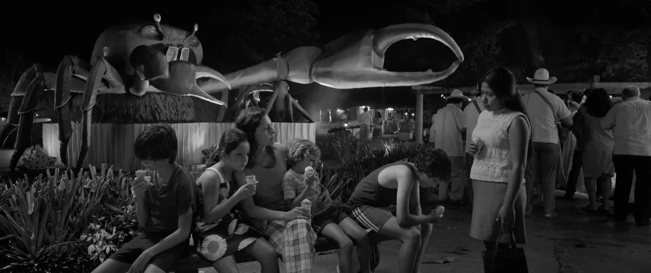 Roma (2018) movie still: wide — A nighttime scene features a large concrete crab statue behind a family on a bench. A woman stands to the right…