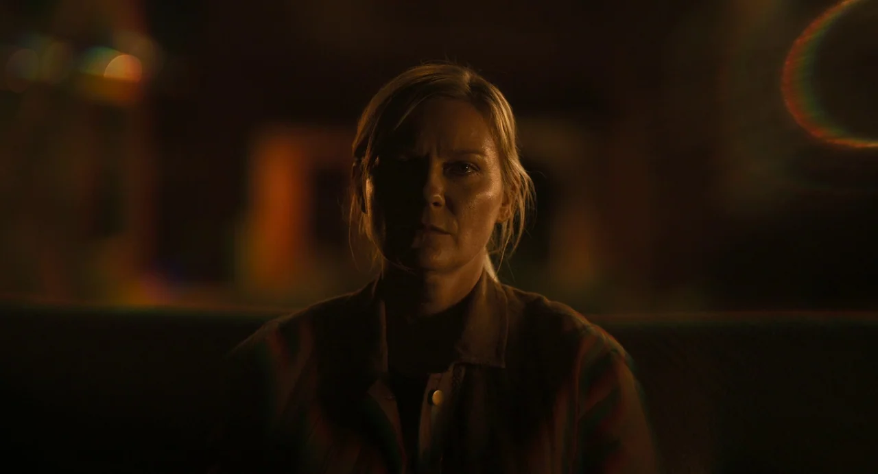 Civil War (2024) movie still: closeup — A dark room with out-of-focus highlights appearing on the periphery. A blonde woman stares into the camera…