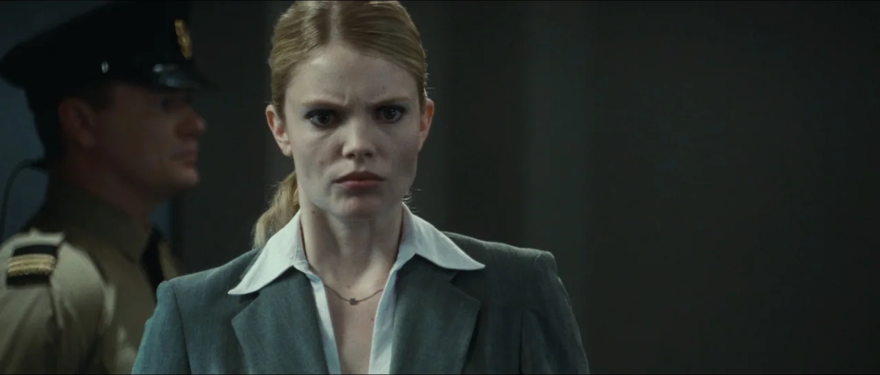 Slackers (2002) movie still: medium — A dimly lit indoor space with neutral-colored walls in a professional setting. A woman in a grey blazer stands…