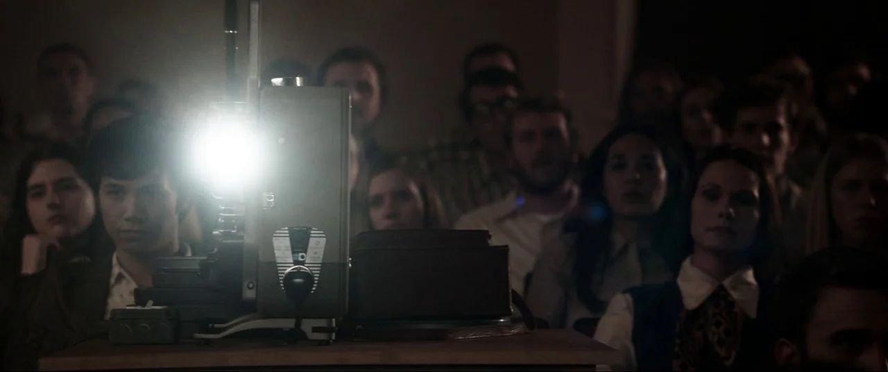 The Conjuring (2013) movie still: medium — A group of people sit in a dark, 1970s-style theater watching a film. A film projector sits prominently…