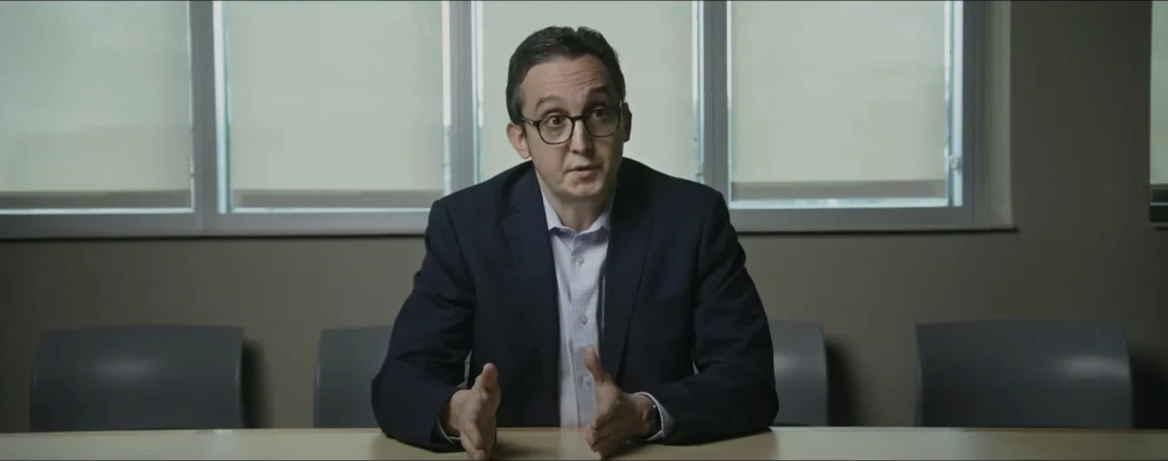 Hunting for Hedonia (2019) movie still: medium — A man sits at a table in a modern office with large window backdrops. He sits facing forward and…
