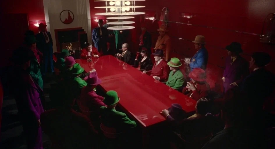 Dick Tracy (1990) movie still: wide — A long red table fills the center of a room with matching red walls. Numerous men in brightly colored suits…