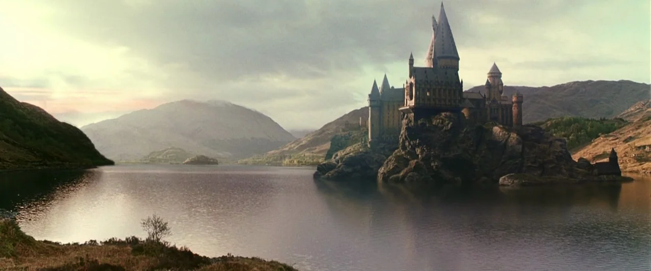A large stone castle sits perched on a rocky cliff overlooking a calm lake. Mountains rise in the background under a soft overcast sky.
