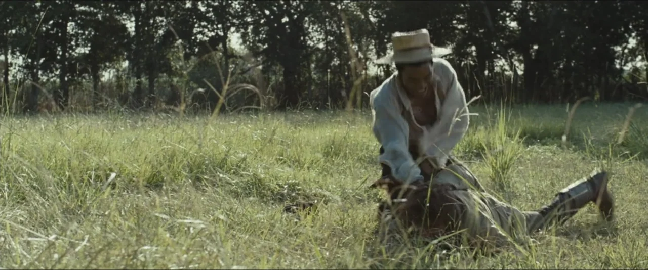 12 Years a Slave (2013) movie still: medium — An open grassy field under trees during the day. One man subdues another man on the ground.