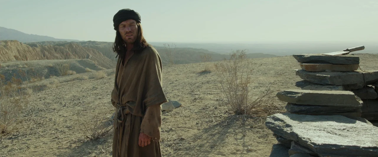 Last Days in the Desert (2015) movie still: fullbody — An arid, rocky desert plateau stretches toward distant hills under a pale sky. A man wearing…