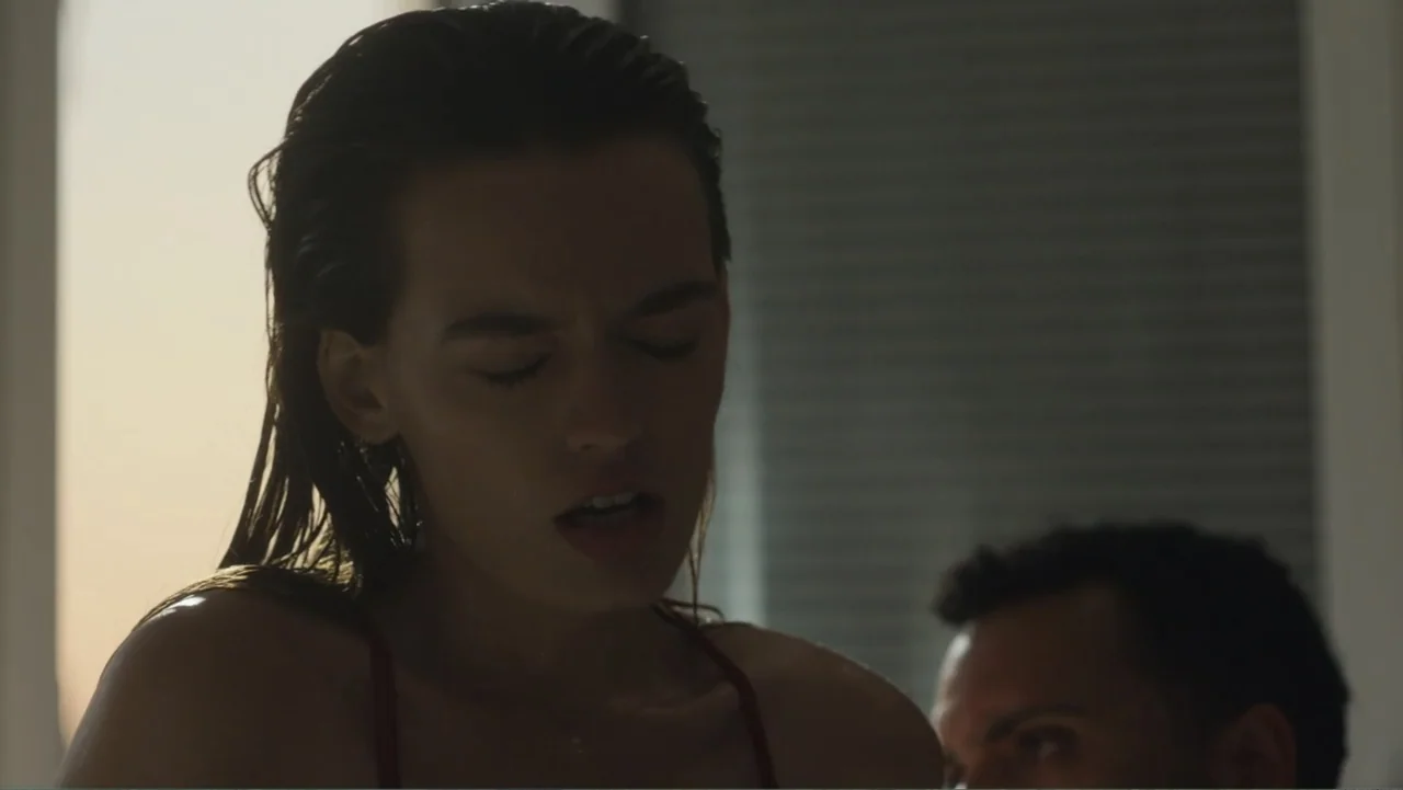 Hot Milk (2025) movie still: medium — A modern room interior with a window showing golden hour light in the background. A woman with wet hair faces…