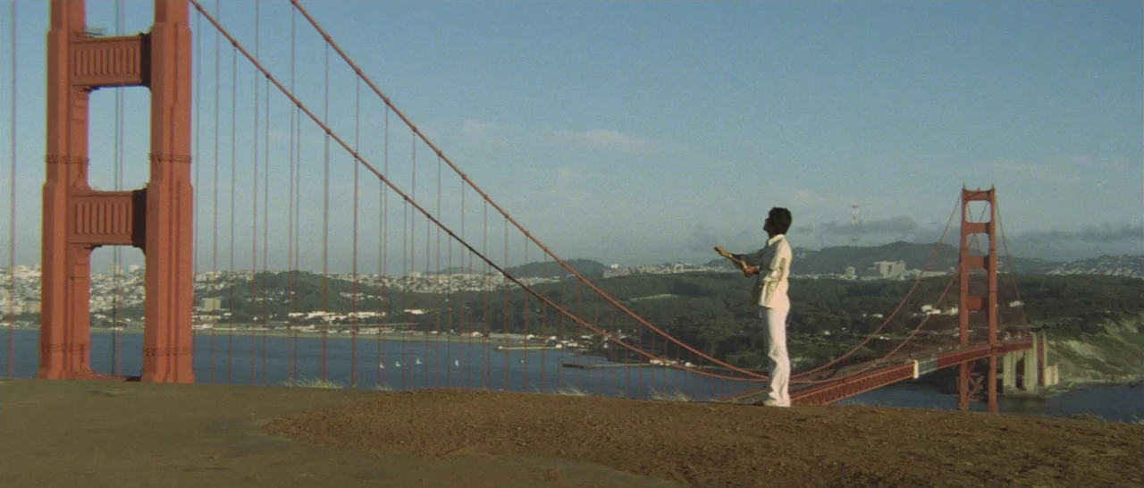Take Me Away! (1978) movie still: wide — The Golden Gate Bridge spans across a bay under a clear, bright blue sky. A man stands on a dirt path in…