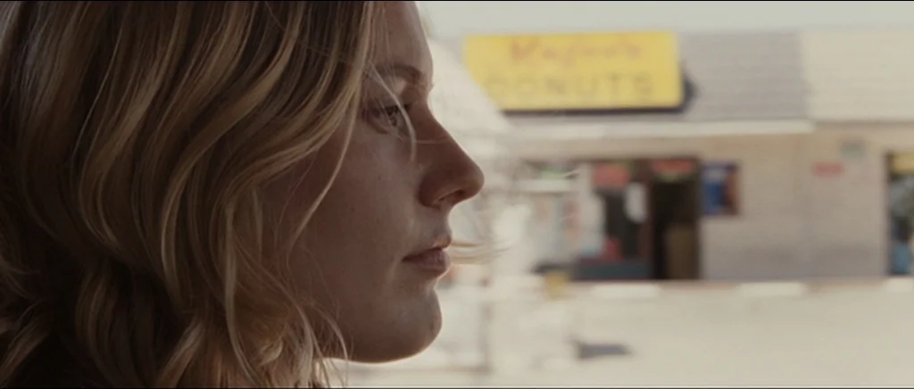Greenberg (2010) movie still: closeup — A sunny parking lot in front of a small commercial storefront with signage. A young blonde woman is centered…