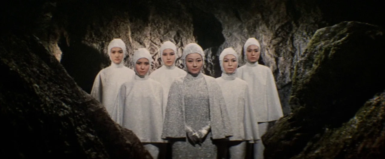 Destroy All Monsters (1968) movie still: medium — Six women stand in a line within a rocky cave formation. They are dressed in identical metallic…