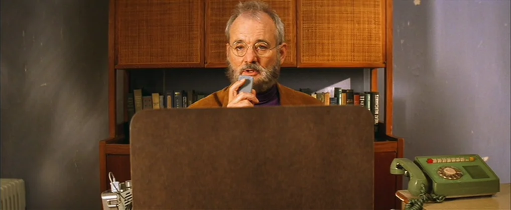 The Royal Tenenbaums (2001) movie still: medium — An office interior features a wooden hutch cabinet filled with books behind the subject. A man in…
