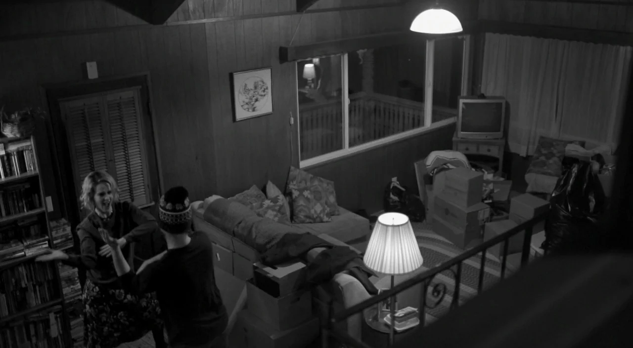 Blue Jay (2016) movie still: wide — A cluttered living room interior features wood-paneled walls and several cardboard moving boxes. A woman and a…