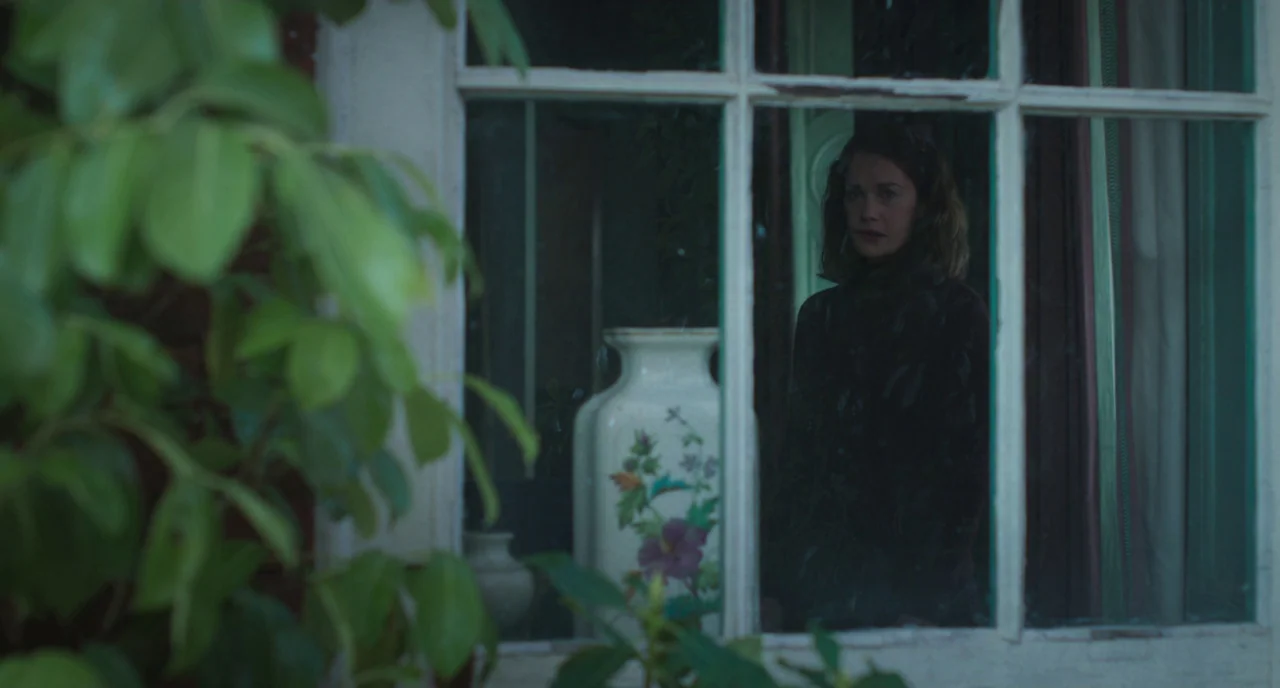 The Little Stranger (2018) movie still: medium — A woman stands behind a multi-paned window, framed by green foliage in the foreground. Beside her…