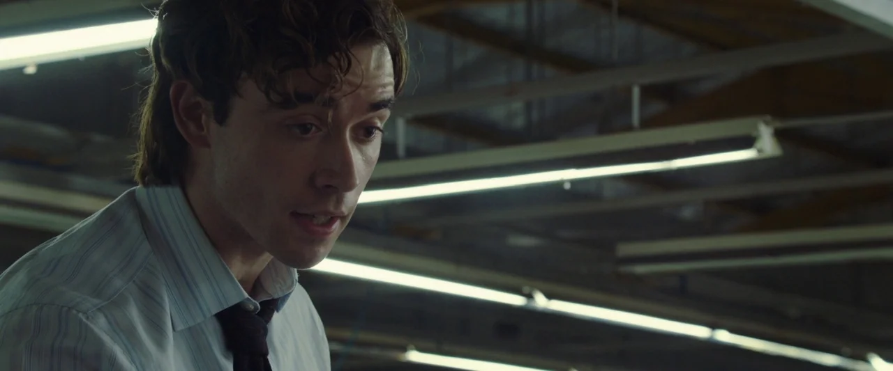 Greed (2019) movie still: medium — A high-ceilinged industrial space with exposed structural beams and overhead lighting fixtures. A young man with…