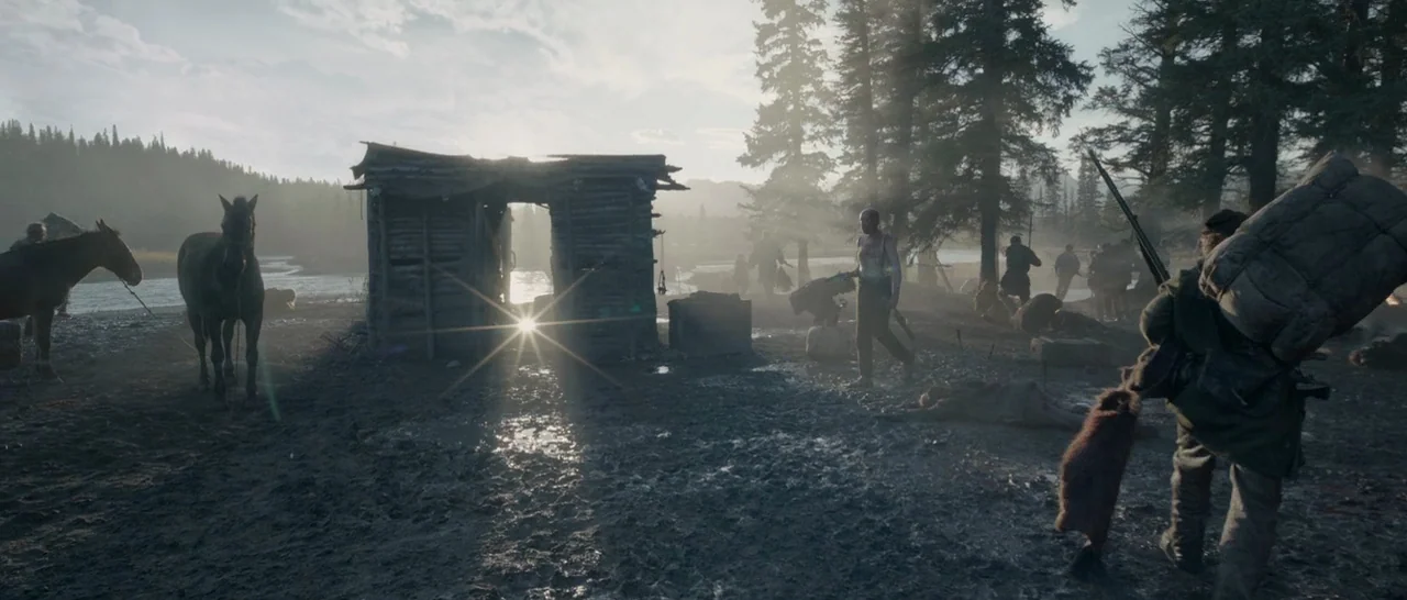 The Revenant (2015) movie still: wide — A rugged riverbank encampment features a small, dilapidated log structure against a wide river. Several men…