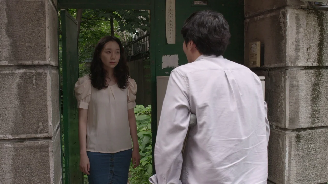 Yourself and Yours (2016) movie still: medium — A woman stands within an open green gate attached to stone pillars. A man with a crutch held under…