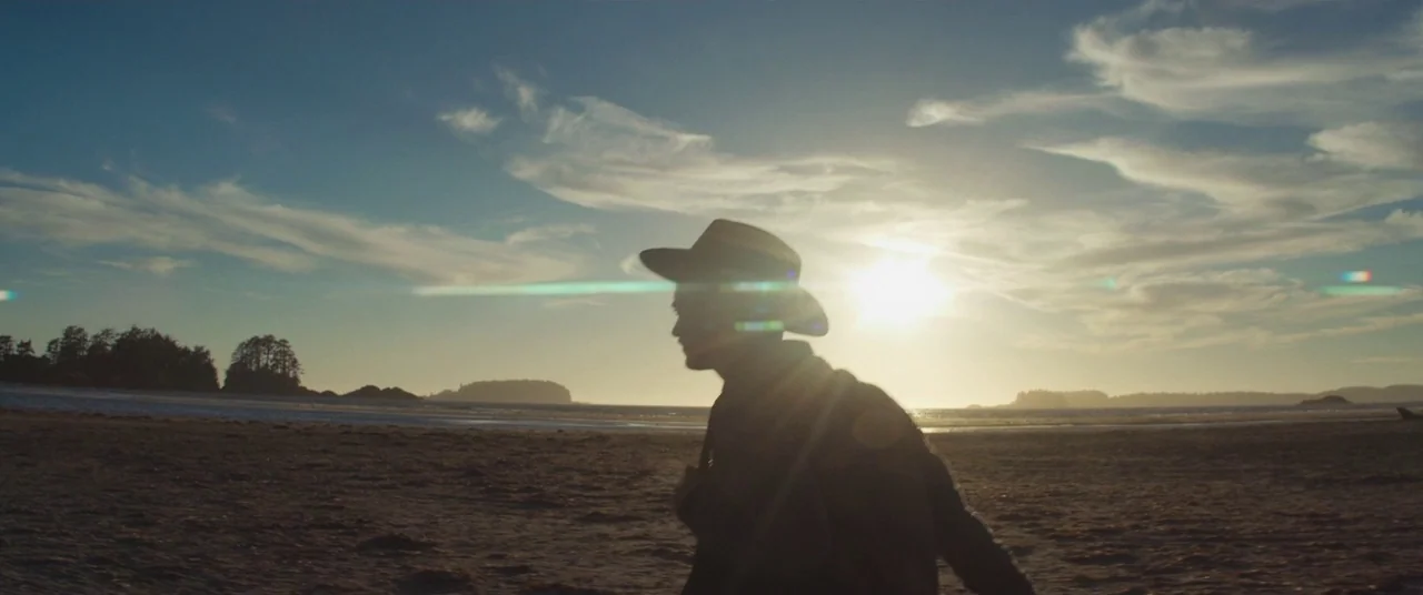 Come To Daddy (2019) movie still: medium — A wide sandy beach stretches toward a distant coastline under a golden sunset sky. A man in a hat walks…