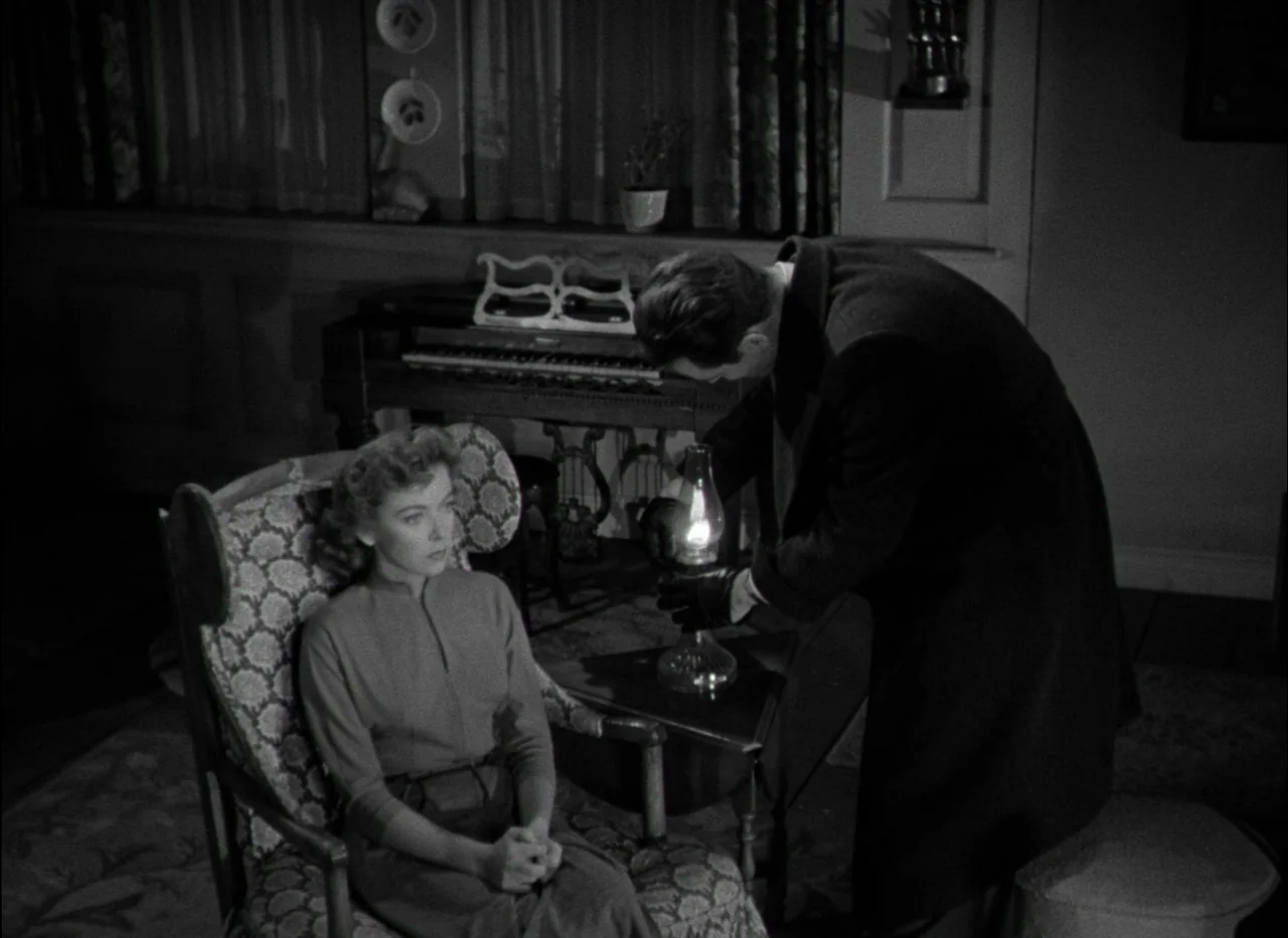 On Dangerous Ground (1951) movie still: medium — A dimly lit room contains an upright piano and a floral-patterned chair. A man in a dark coat…