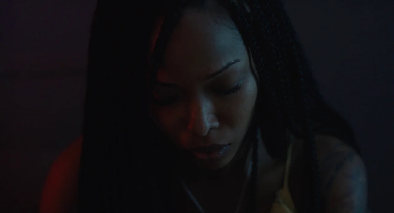 Test Pattern (2019) movie still: closeup — A dimly lit interior space with shadows cast across the subject. A young woman with braids looks downward…