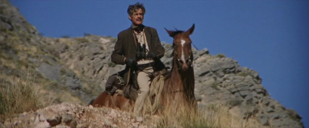 The Wild Bunch (1969) movie still: fullbody — A man on horseback traverses a steep, arid mountain slope under a cloudless sky. He carries binoculars…
