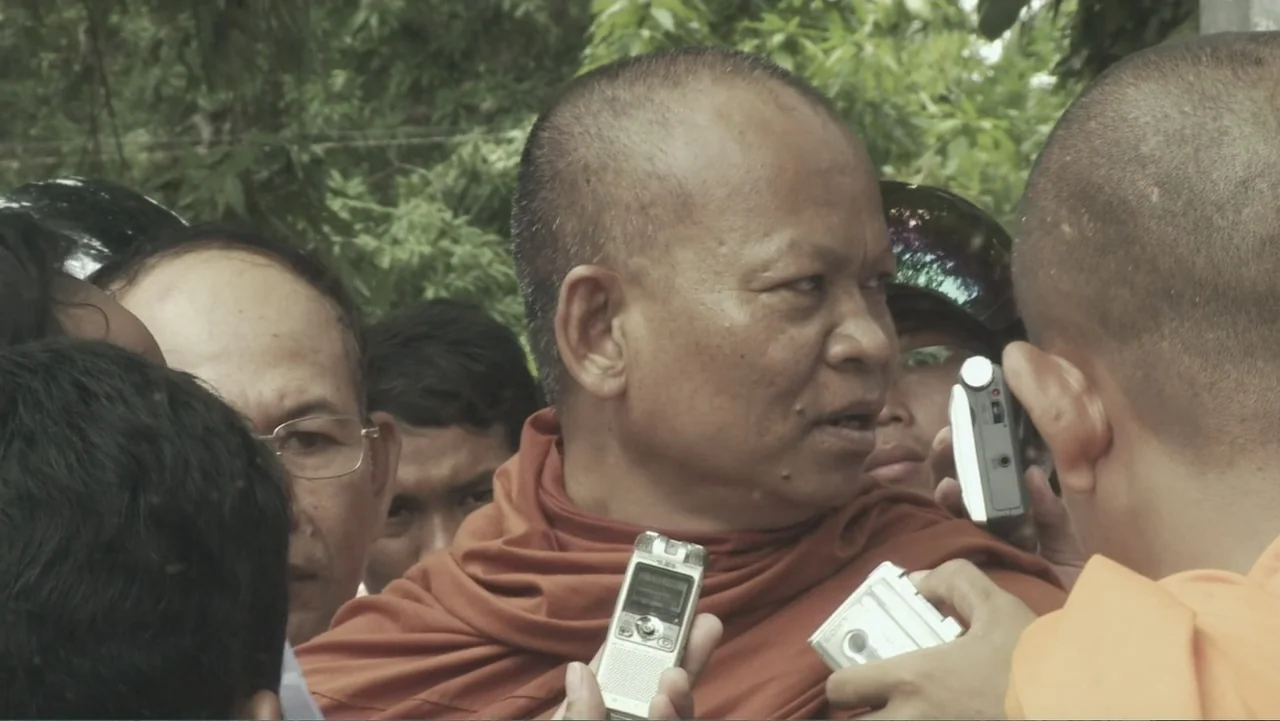 A Cambodian Spring (2018) movie still: medium — A group of people surrounds an older Buddhist monk in orange robes outdoors. Several hands hold…