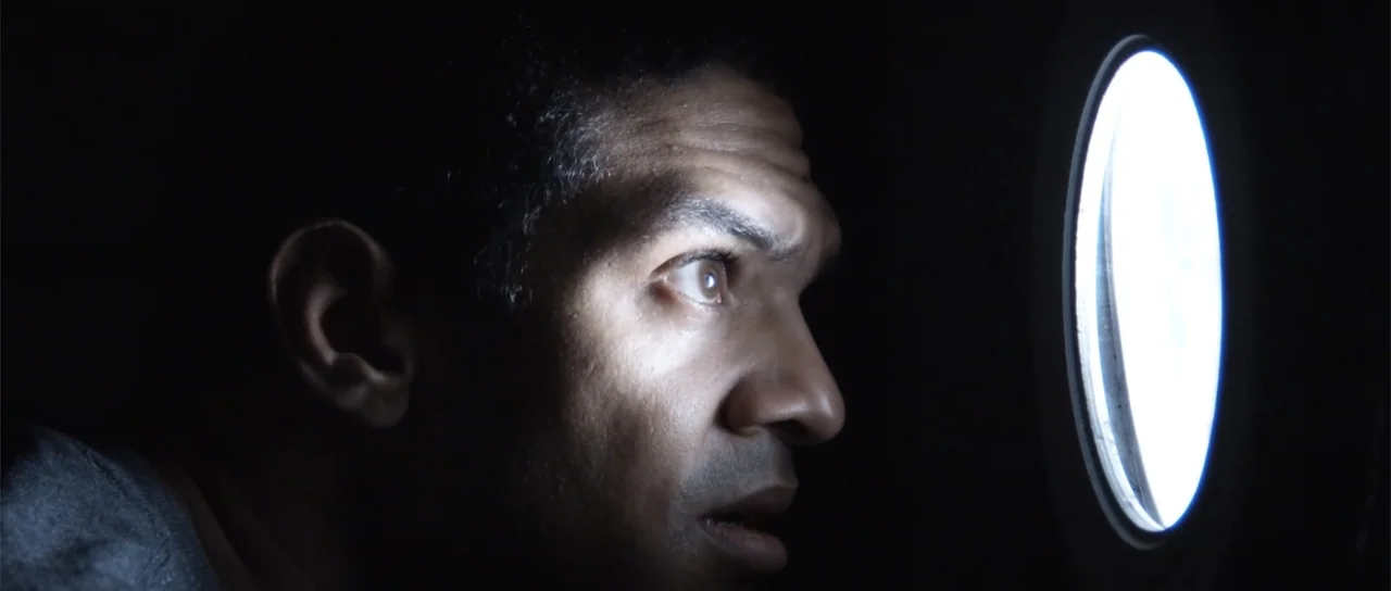Astronaut: The Last Push (2012) movie still: closeup — A dark, cramped interior space features a glowing circular porthole. A man looks through the…