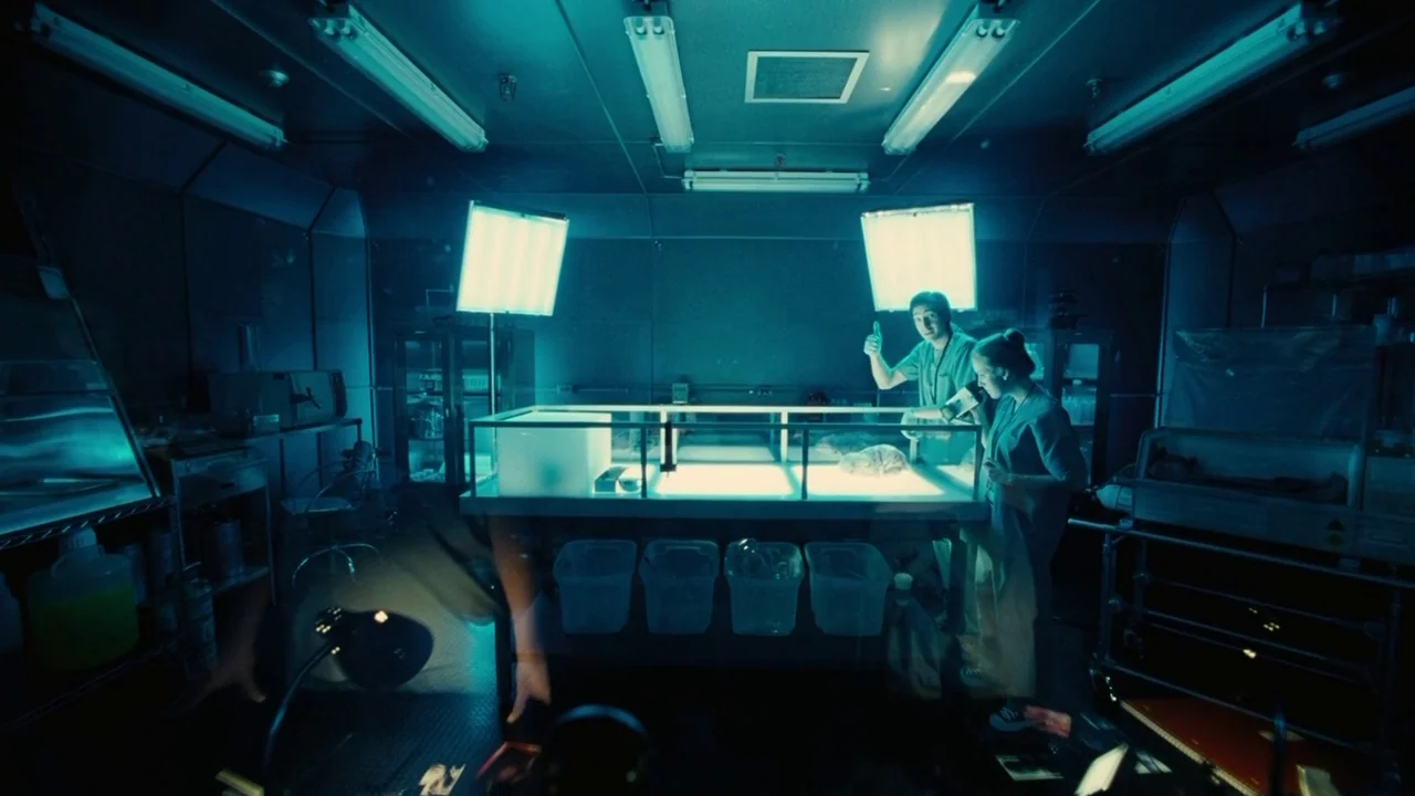 Splice (2009) movie still: wide — A dimly lit sterile laboratory features a central stainless steel workstation with bright illumination. Two…