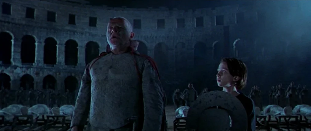 Titus (1999) movie still: fullbody — An expansive ancient stone amphitheater is filled with rows of draped figures at night. A mature man in a cloak…