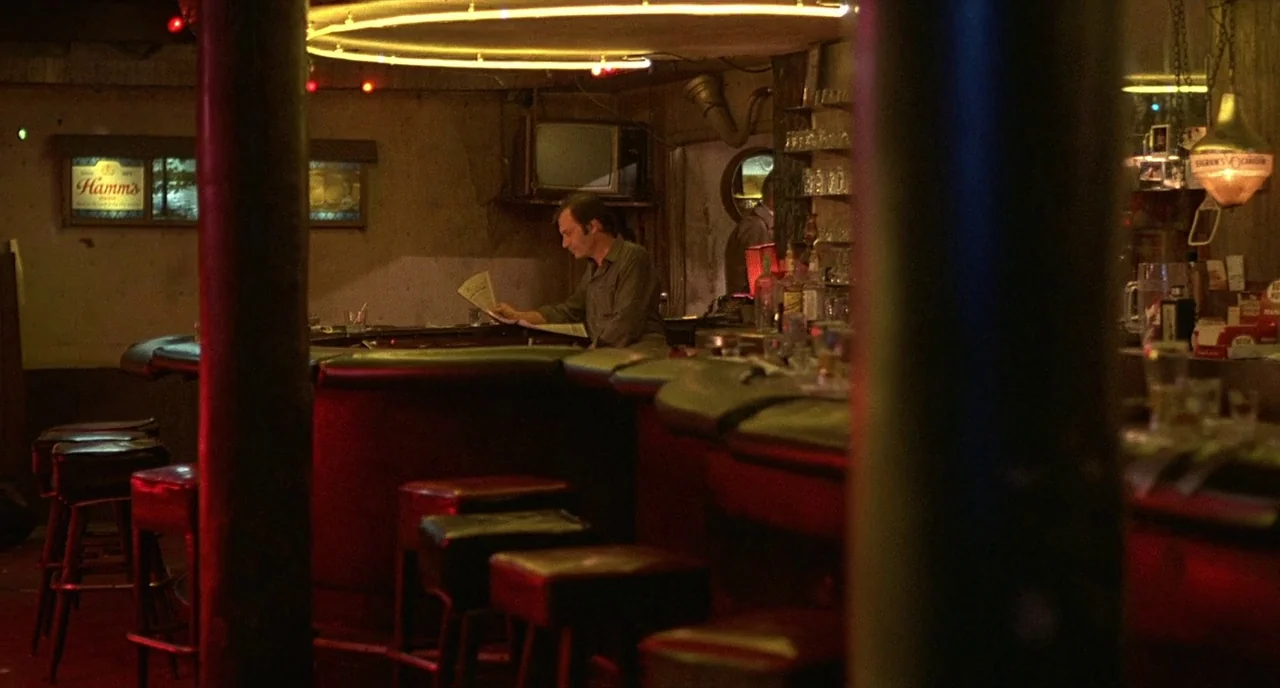 Barfly (1987) movie still: wide — A dark tavern interior features a curved counter and rows of empty stools. A lone man in a dark shirt sits at the…