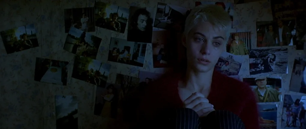 I Want You (1998) movie still: medium — A dark, dimly lit wall is covered with numerous small, candid color photographs. A young woman with a short…