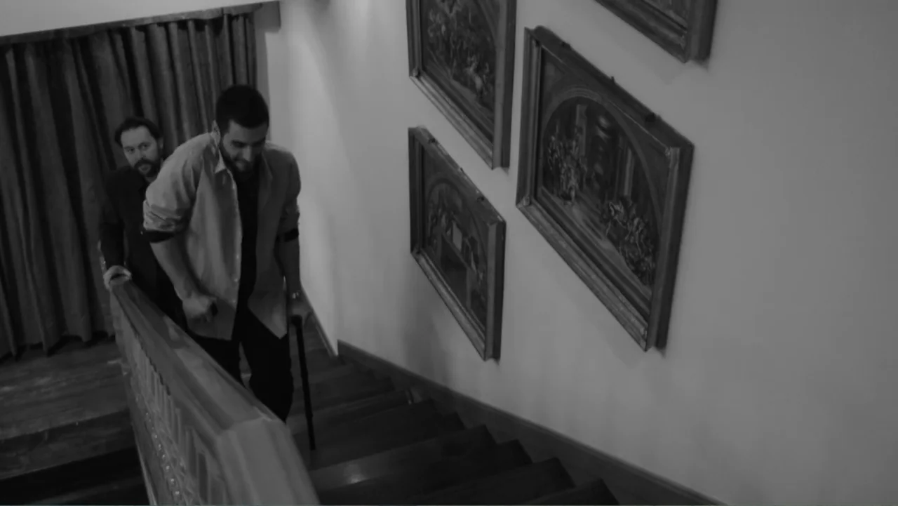 Creditors (2025) movie still: medium — A wooden staircase in a house with framed paintings hanging on the wall. A man using a crutch walks up the…