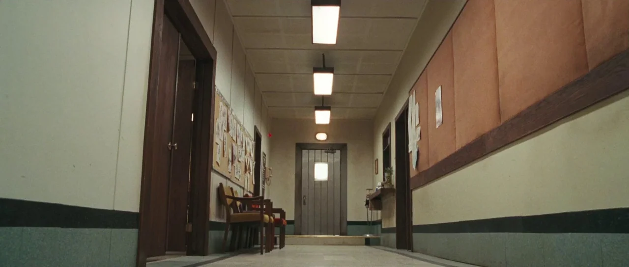 Surveillance (2008) movie still: establishing — A long, symmetrical hallway features paneled walls and multiple doors under a dropped ceiling. Empty…