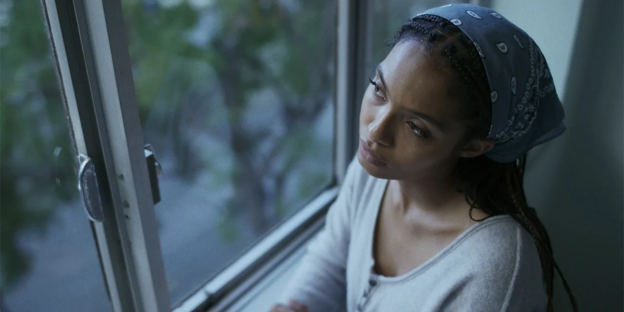 Sitting in Bars with Cake (2023) movie still: medium — A woman stands indoors near a window during daylight hours. She is looking outside with a…
