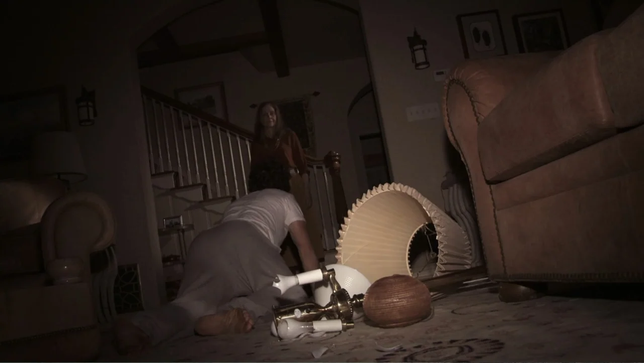 Paranormal Activity: The Ghost Dimension (2015) movie still: medium — A dark living room contains a carpeted floor, a couch, and a staircase. A man…