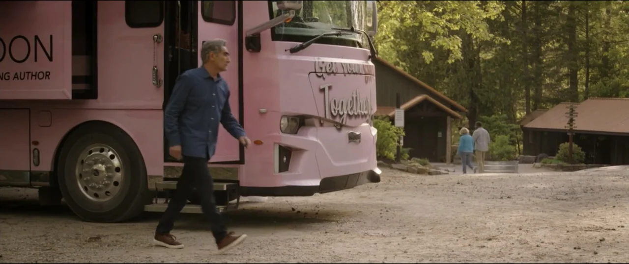 Summer Camp (2024) movie still: fullbody — A gravel parking area sits in front of wooden buildings and dense forest trees. An older man in a blue…