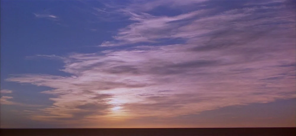 Lawrence Of Arabia movie still: establishing — A vast horizon separates the dark earth from a wide, cloud-filled sky. wispy clouds drift across the…